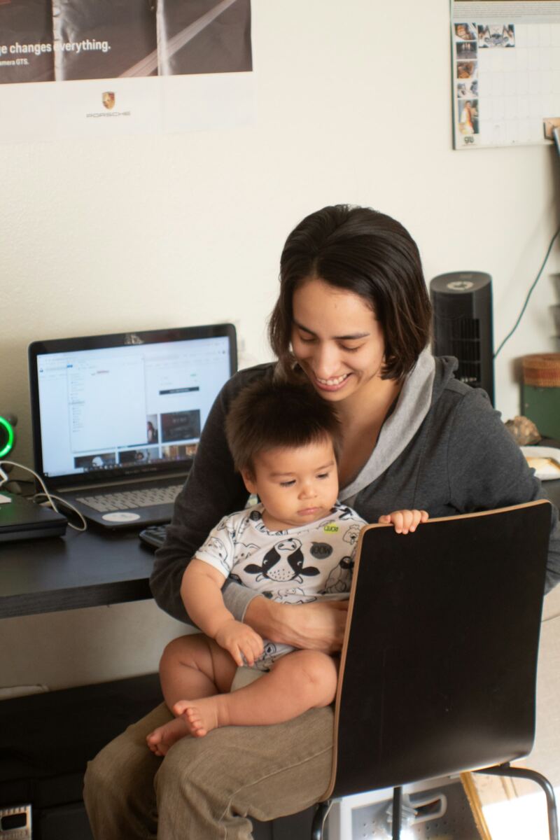 A parent working at a laptop while holding a baby